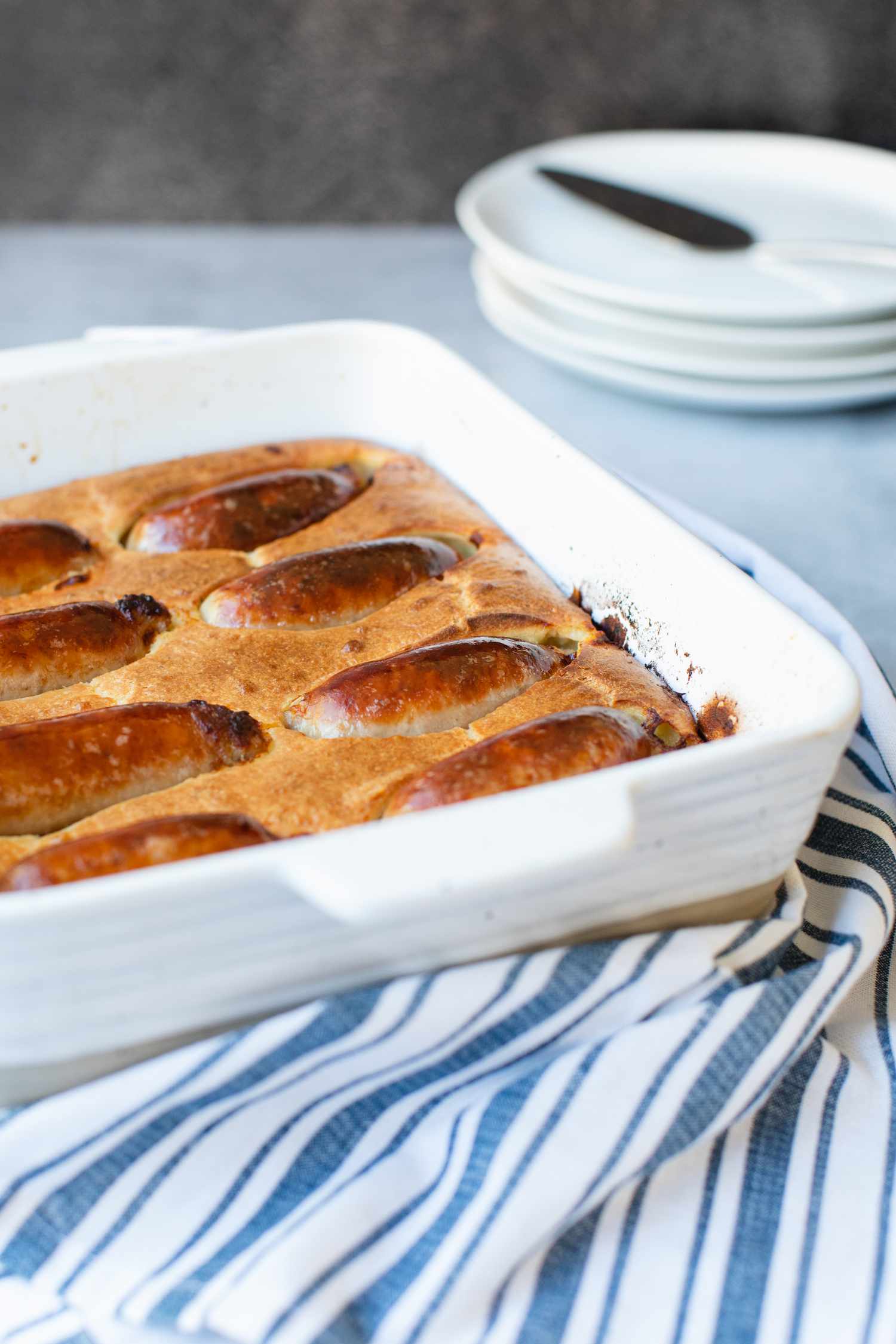Toad in the hole in a white baking dish with a blue striped linen and stack of plates around it.