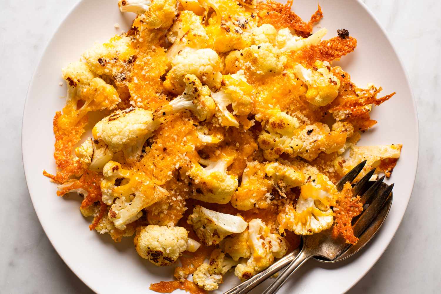 A plate of roasted cauliflower topped with melted cheese and breadcrumbs served with a fork and spoon on the side