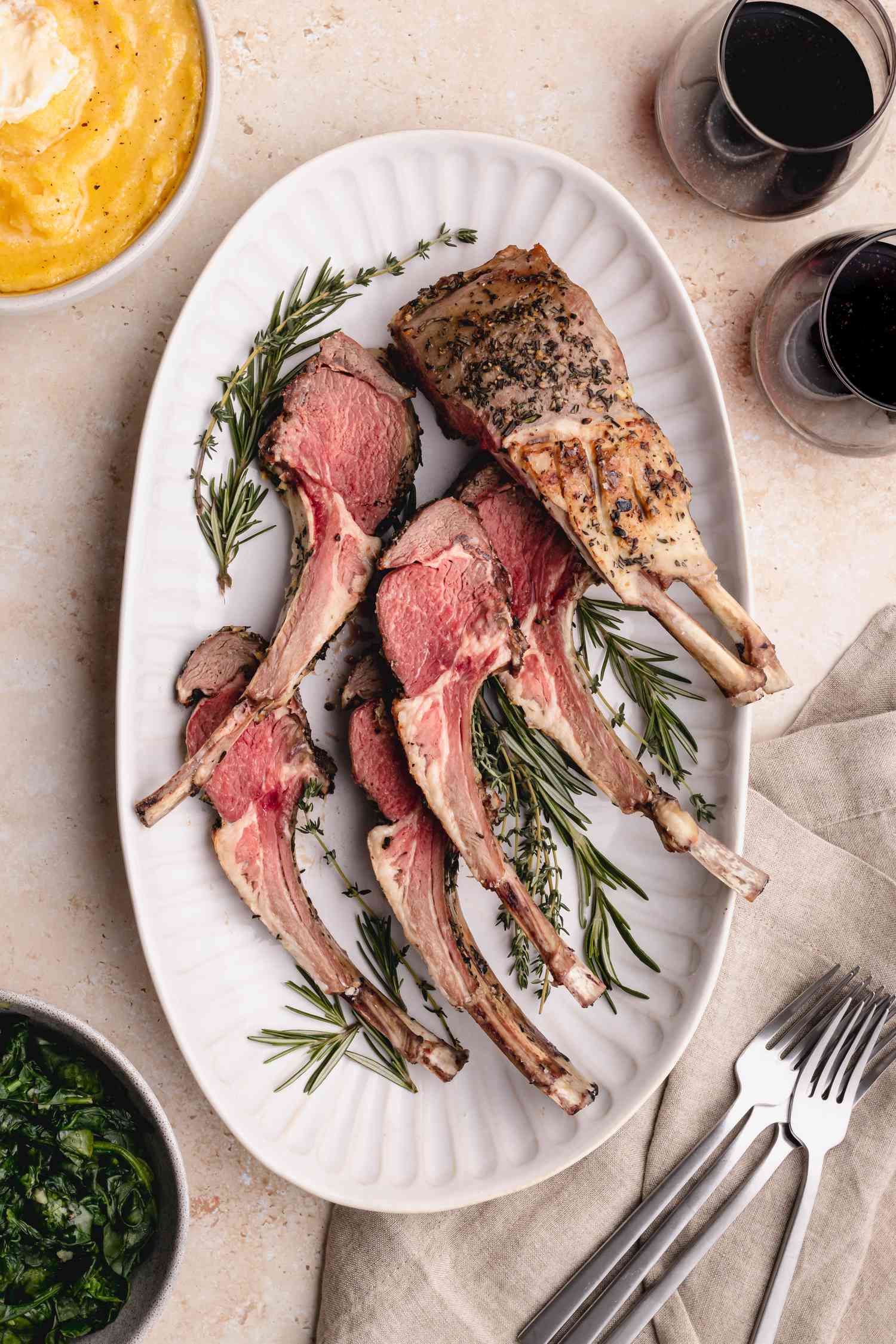 Rack of lamb cut into chops on white platter with 2 glasses of wine and utensils