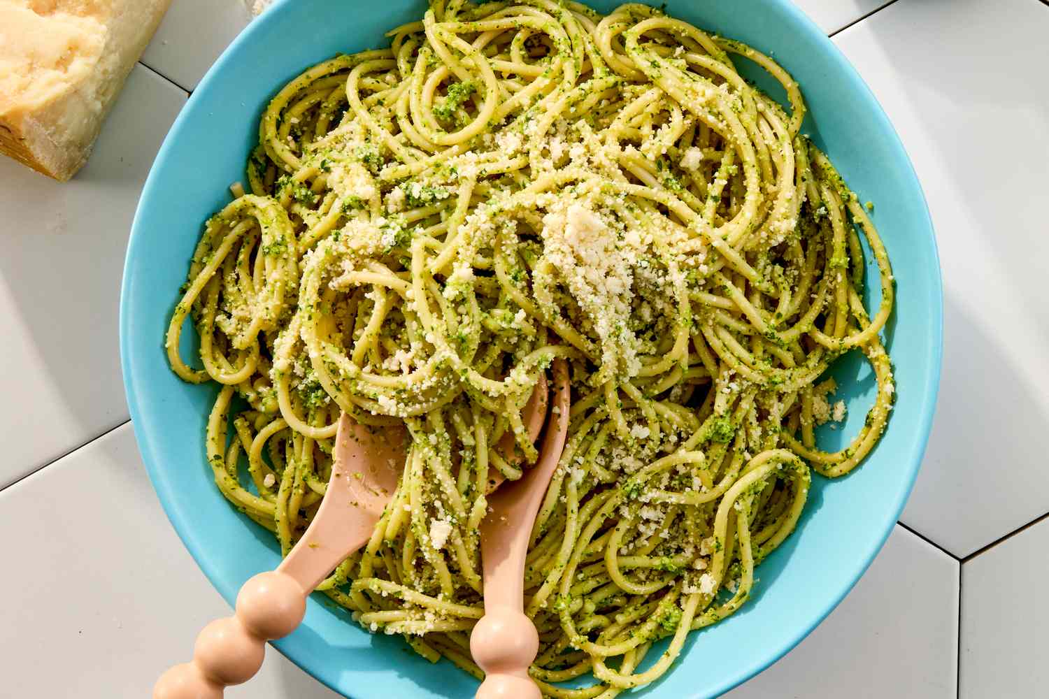 A bowl of pasta coated with pesto garnished with grated cheese with wooden utensils placed on top