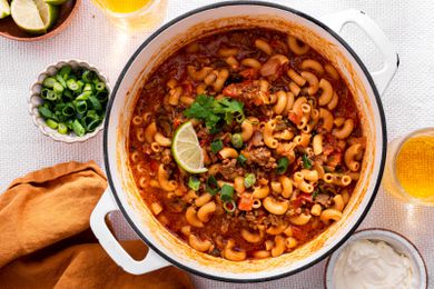 One-Pot Taco Pasta in a Pot, and in the Surroundings, a Bowl With Lime Wedges, a Bowl With Sliced Green Onions, a Bowl With Sour Cream, Glasses With a Drink, and a Mustard Colored Table Napkin