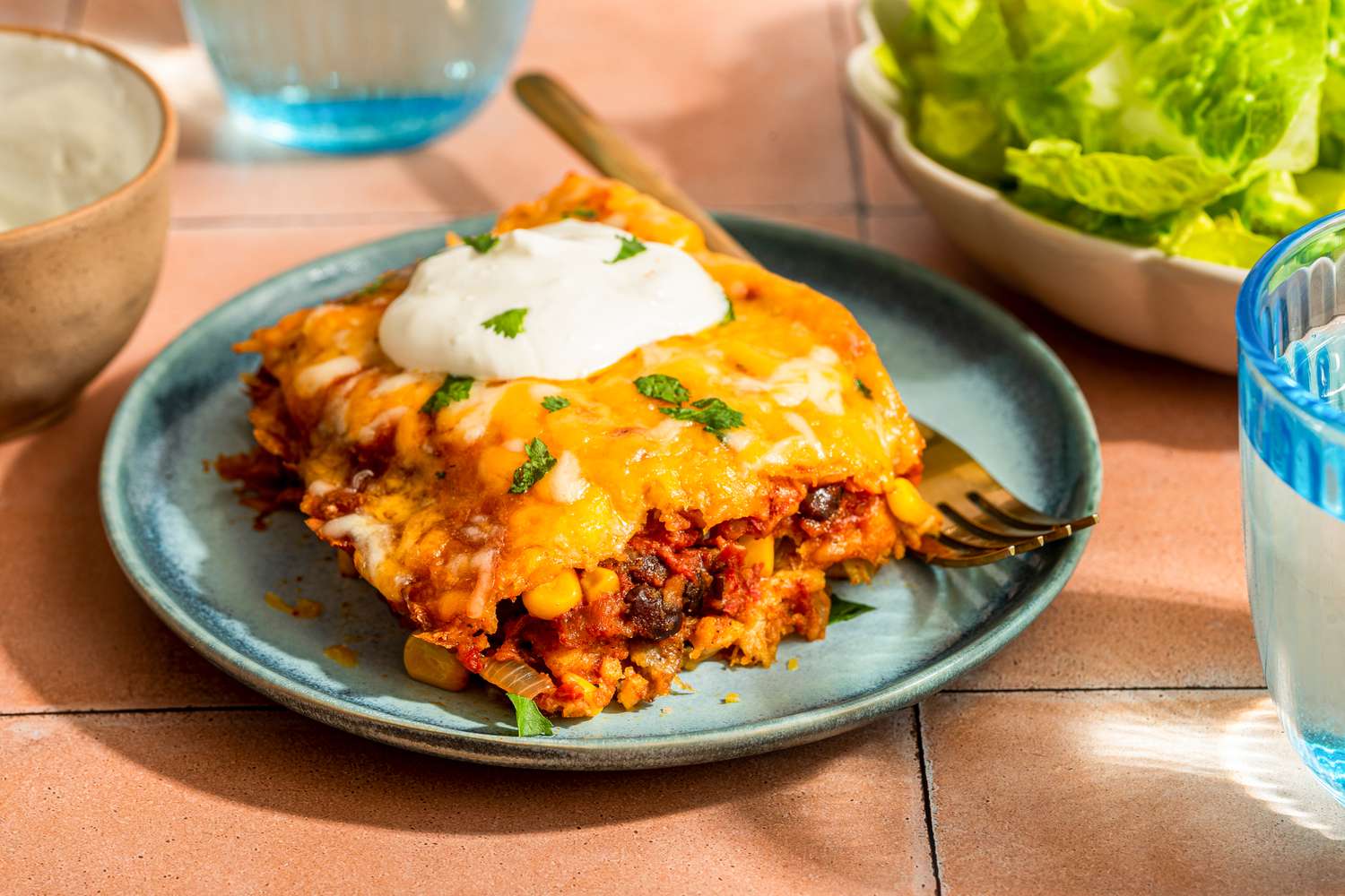 Serving of chicken taco casserole on a plate topped with a dollop of sour cream at a table setting with glasses of water, a bowl of side salad, and a bowl with more sour cream