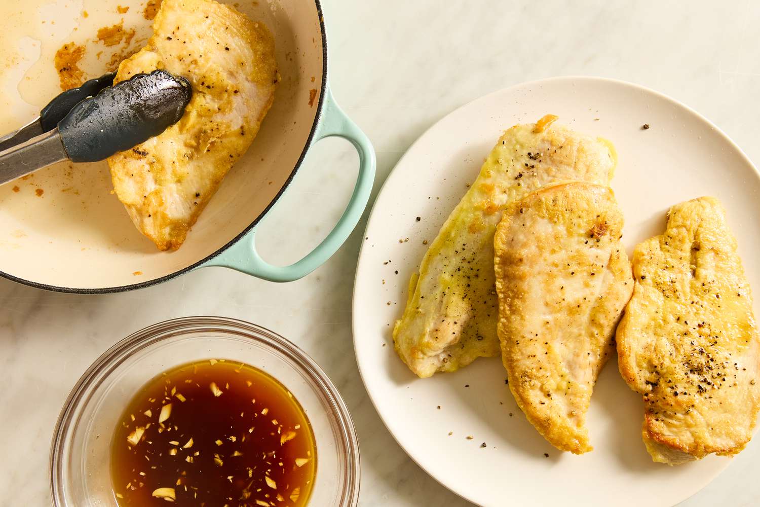 Overhead view of tongs removing a chicken breast from a dutch oven next to a white plate of three chicken breasts and a bowl of the soy sauce mixture for Honey Soy Chicken recipe