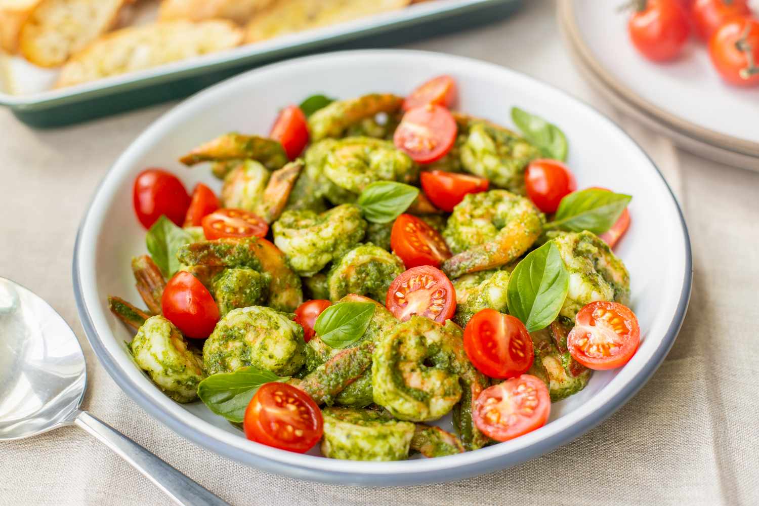 5-Ingredient Pesto Shrimp with Cherry Tomatoes in a Bowl, and in the Background, a Tray of Garlic Bread and Cherry Tomatoes
