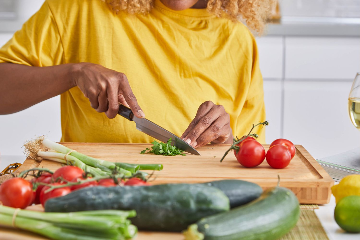 A person wearing a yellow shirt cuts a small bundle of parsley on a cutting board on a mat, and on the board with the parsley, tomatoes and green onions. In on the counter around the cutting board, a glass of wine and more produce (cucumbers, zucchini, tomatoes on a vine, limes and lemons, and green onions.