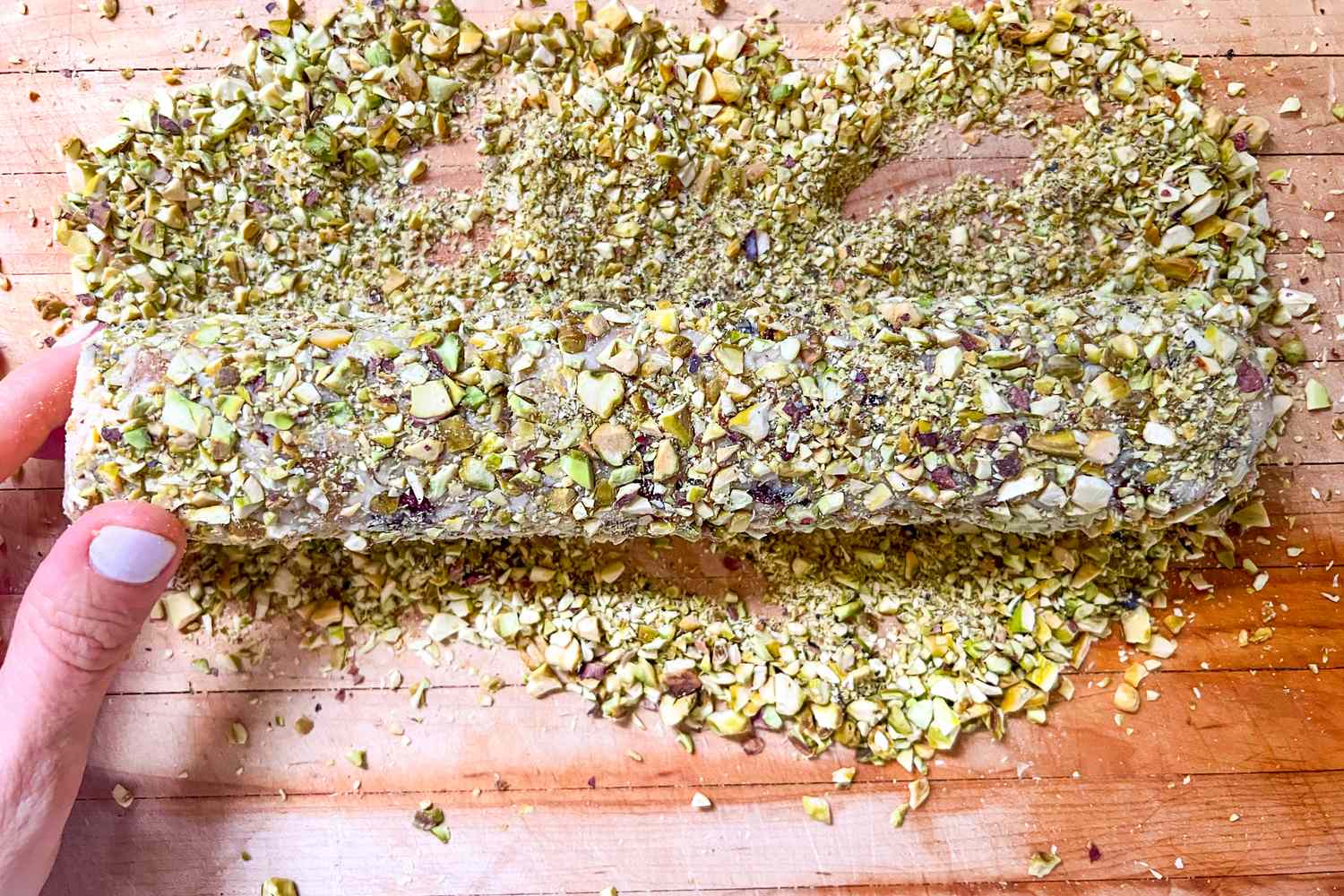 Rolling a dough log in chopped pistachios on a wooden surface