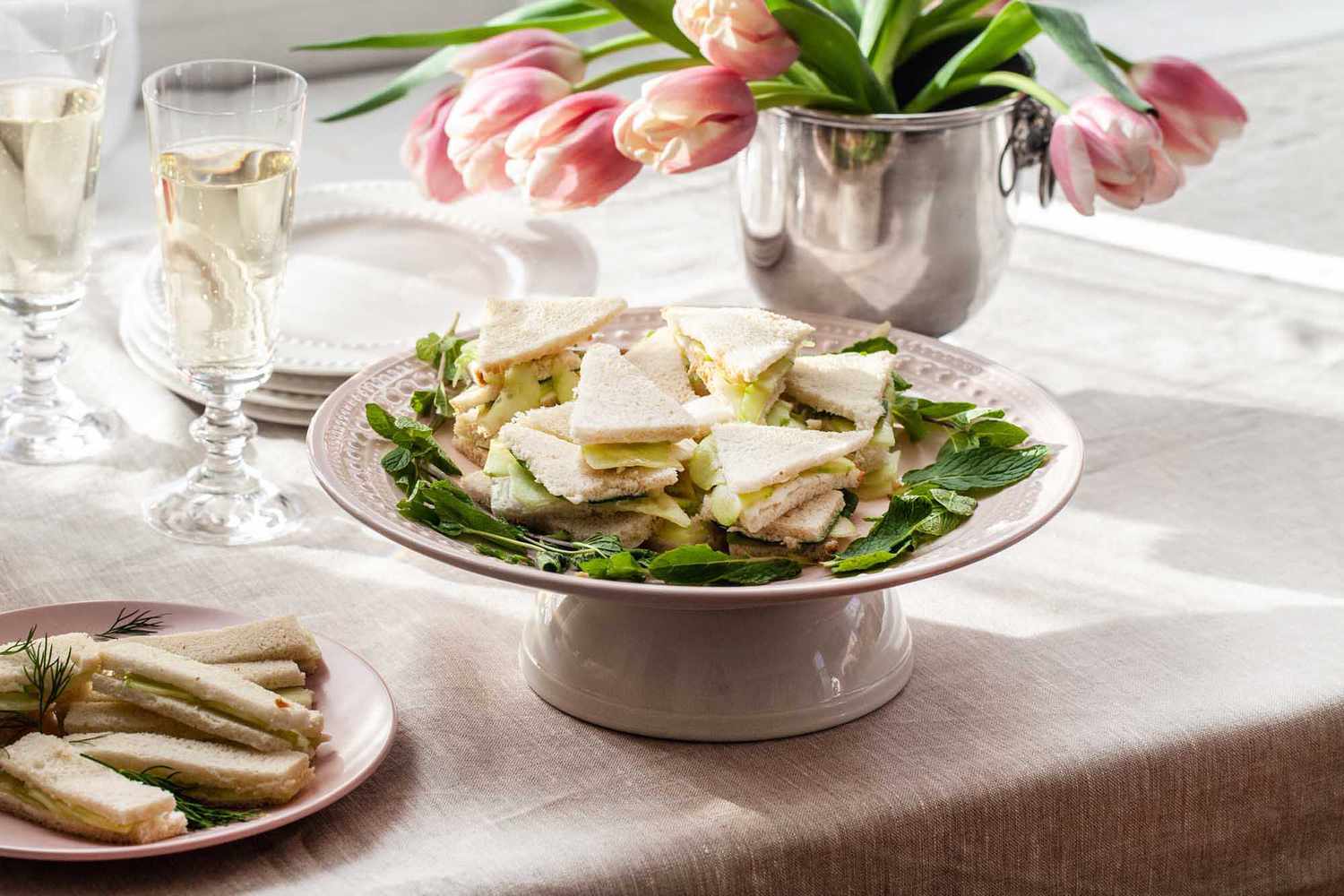 Table set with tulips, champagne and a cake plate with cucumber finger sandwiches on top.