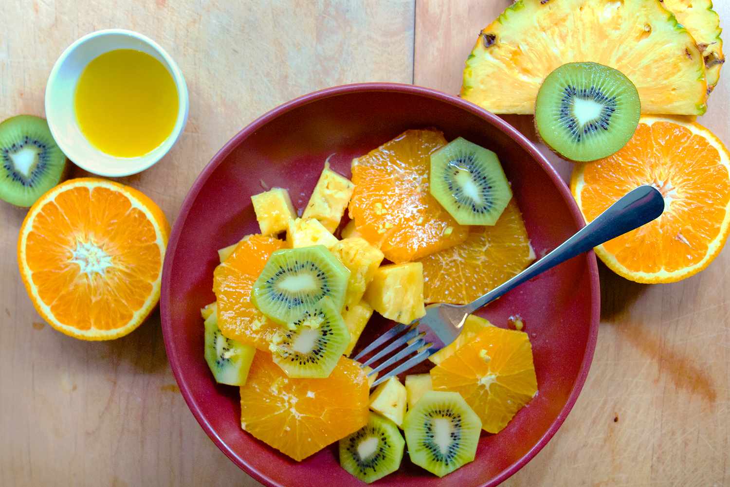 Bowl of fruit salad with pineapple, orange, and kiwi slices, fork in bowl, orange half, and juice on the side