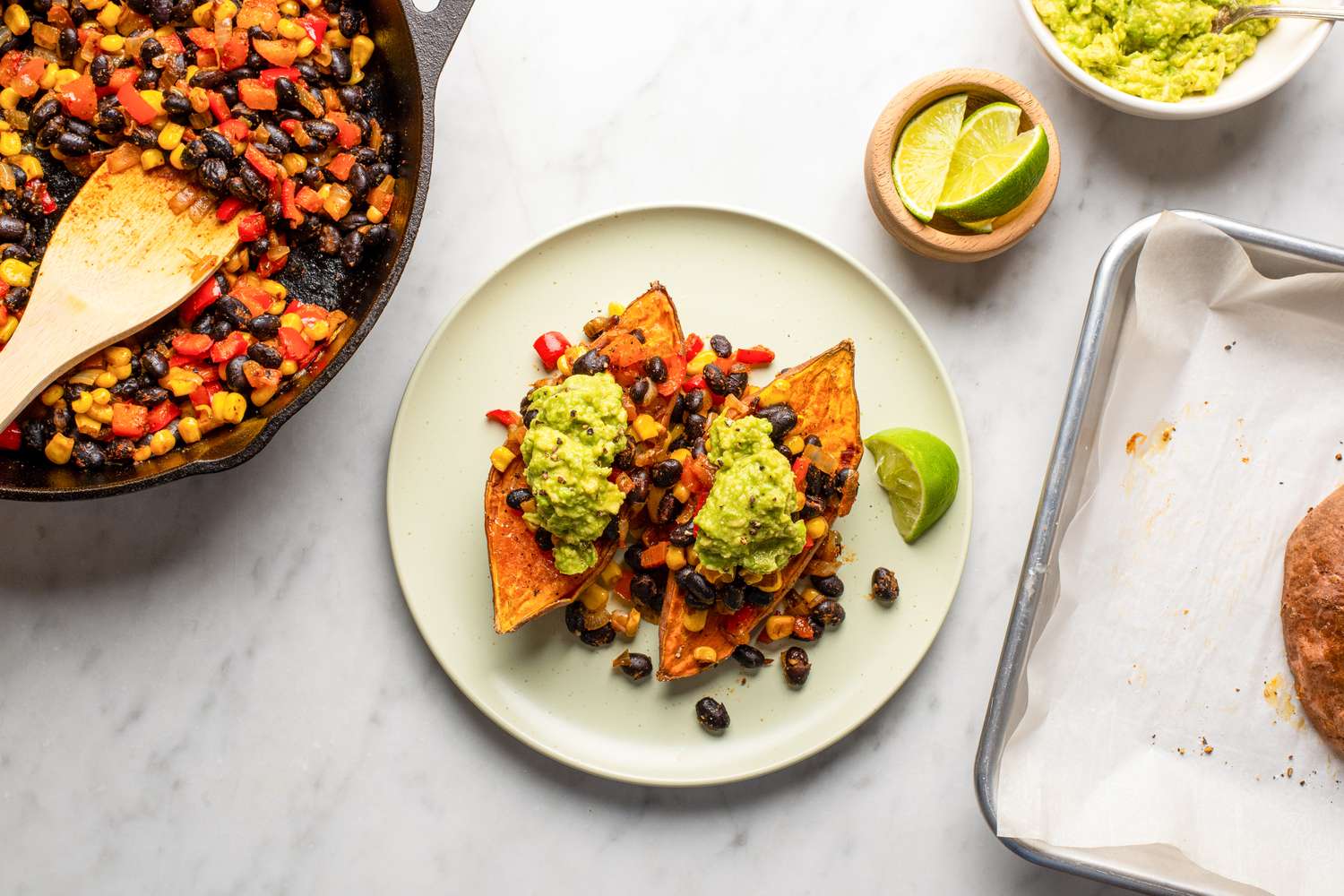 A plate with sweet potato halves topped with black beans and guacamole next to a skillet containing the mixture and a lime garnish beside it