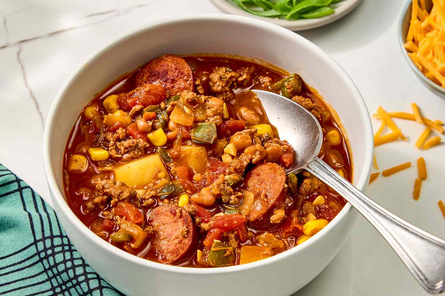Overhead view of a bowl of cowboy stew with a spoon next to a bowl of shredded cheddar, scallions, and a cloth napkin all on a marble countertop