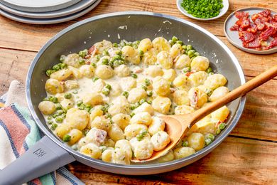 Creamy Skillet Gnocchi With Bacon and Peas in a skillet with a spoon at a table setting with a stack of plates, a bowl of chopped chives, and a small plate of bacon pieces