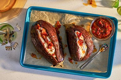 Two whole roasted eggplant on a baking sheet, topped with yogurt and chili crisp, a bowl of chili crisp alongside