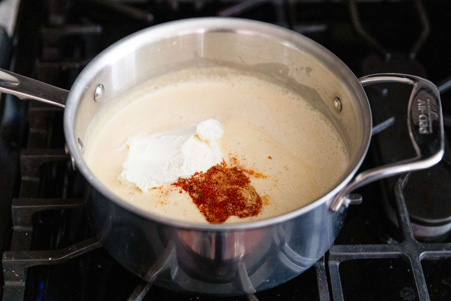 Angled view of a saucepan after adding cream cheese, seasoned salt, garlic powder, paprika, cayenne pepper, turmeric (if using), and mustard simmering on a gas stovetop from Baked Mac and Cheese recipe