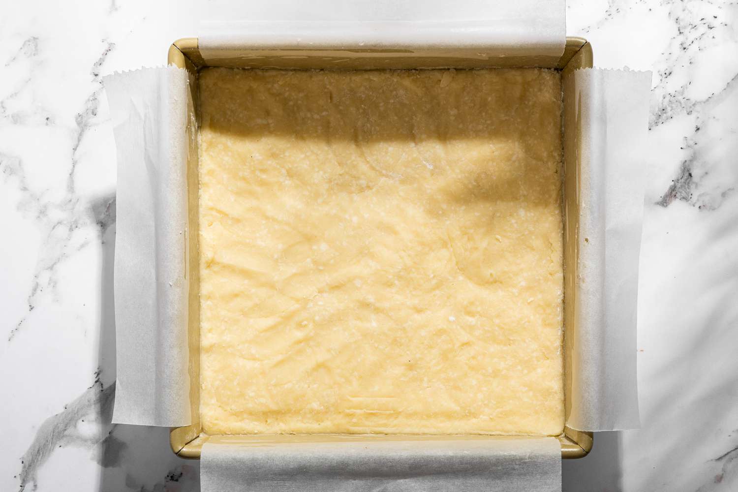 Overhead view of a baking pan with the flour, butter, and powdered sugar mixture pressed into parchment paper for Millionaire Shortbread recipe 