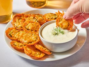 A plate of crispy fried pickles with a hand dipping one into a bowl of white sauce, garnished with a sprig of dill