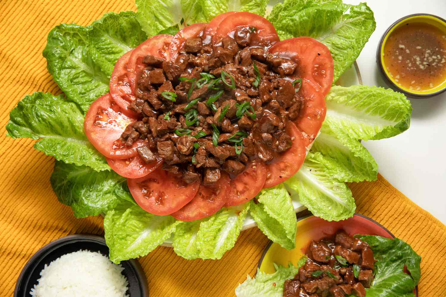 Platter of Lok Lak Served Over Sliced Tomatoes and Lettuce at a Table Setting With a Bowl of Rice, a Plate With a Serving of Lok Lak, and a Bowl of Pepper-lime Sauce 