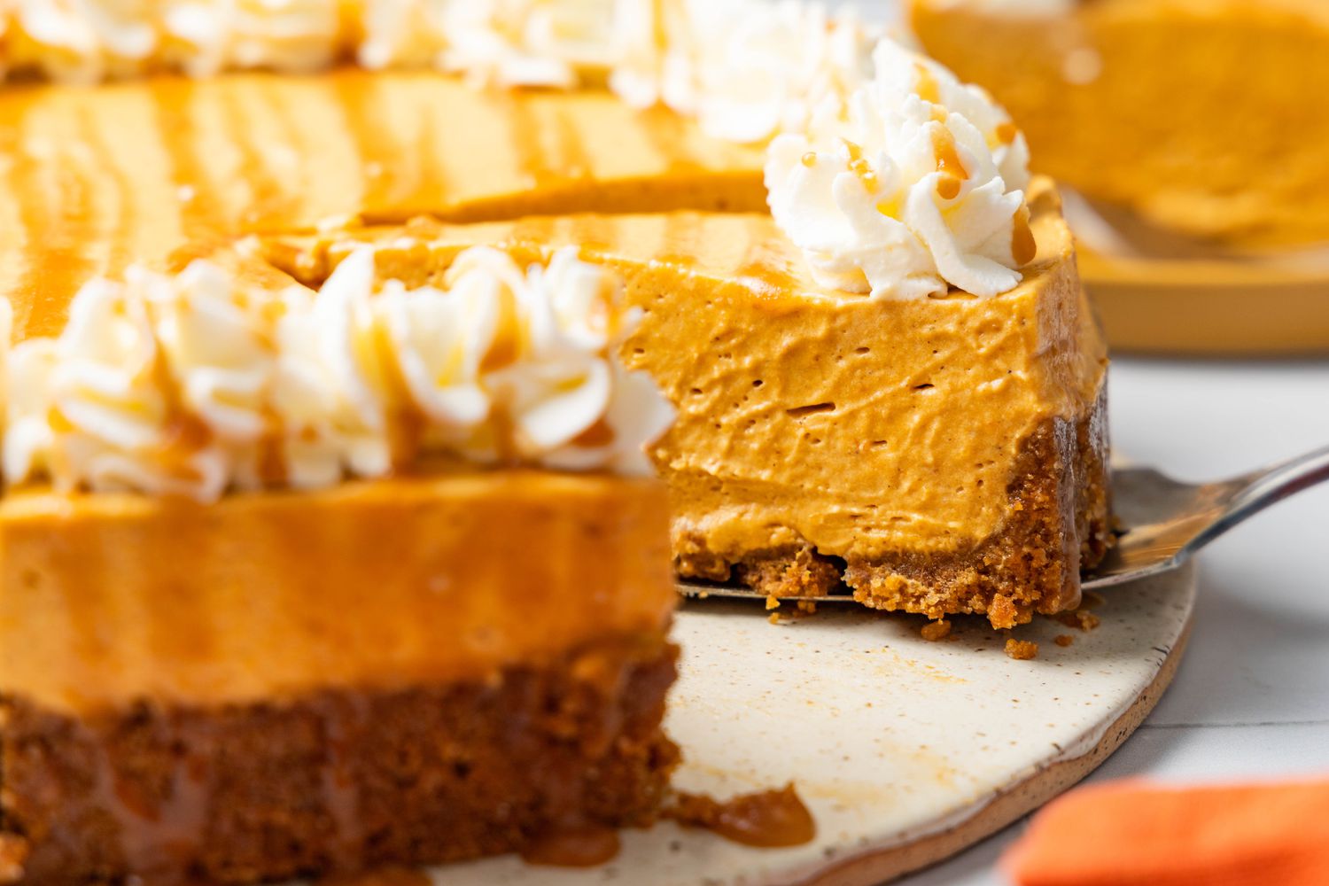 Slice of no-bake cheesecake slightly lifted from platter with the rest, using a pie server, and in the surroundings, an orange kitchen towel and a plate with a slice of cheesecake