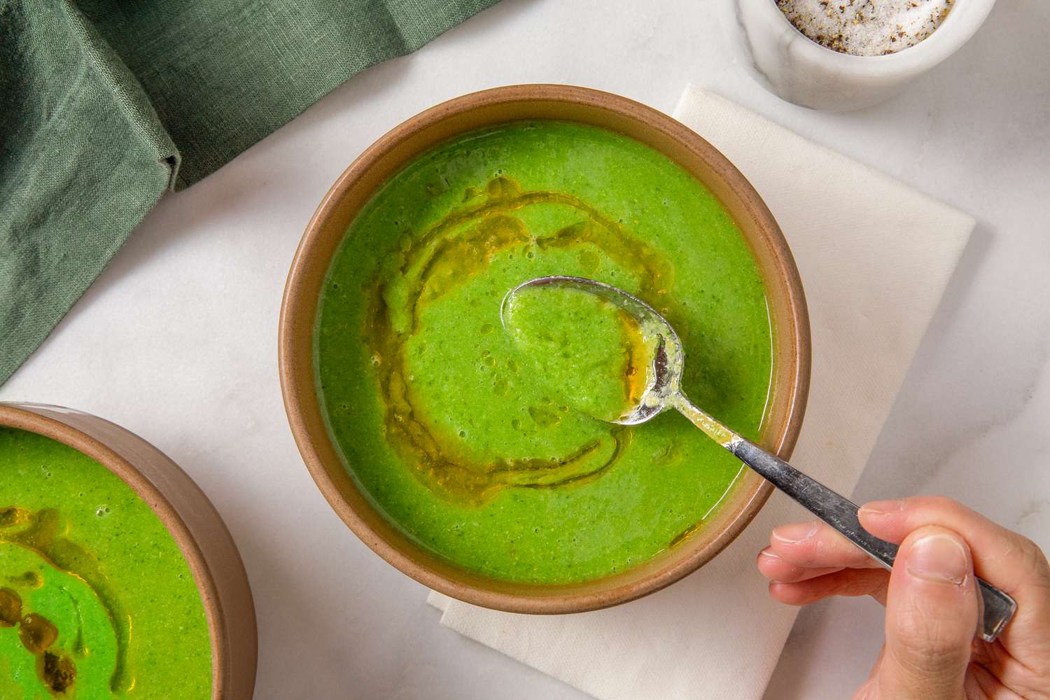 A bowl of broccoli soup with a hand scooping some of the soup out with a spoon