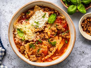 Lasagna Soup in a Bowl With Noodles, Sausage, Ricotta, and Fresh Basil, and in the Surroundings, Another Bowl With Crushed Red Peppers, a Saucer With Basil Leaves, and a Blue and White Checkered Kitchen Towel 