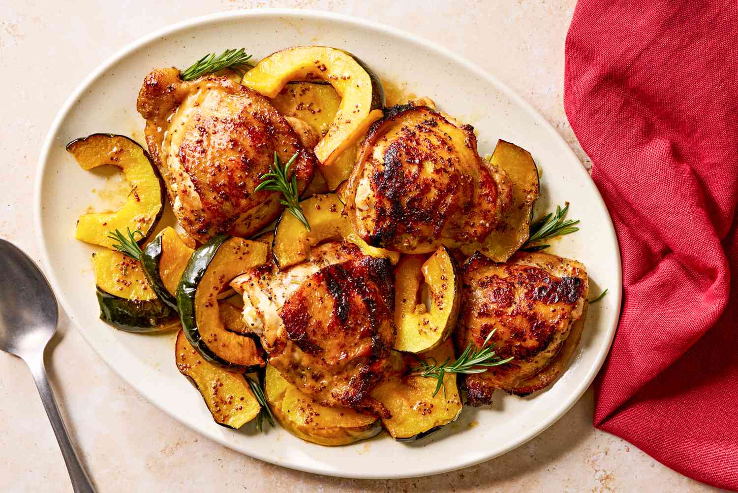 Overhead view of a white platter of maple glazed chicken and squash next to a spoon and red cloth napkin