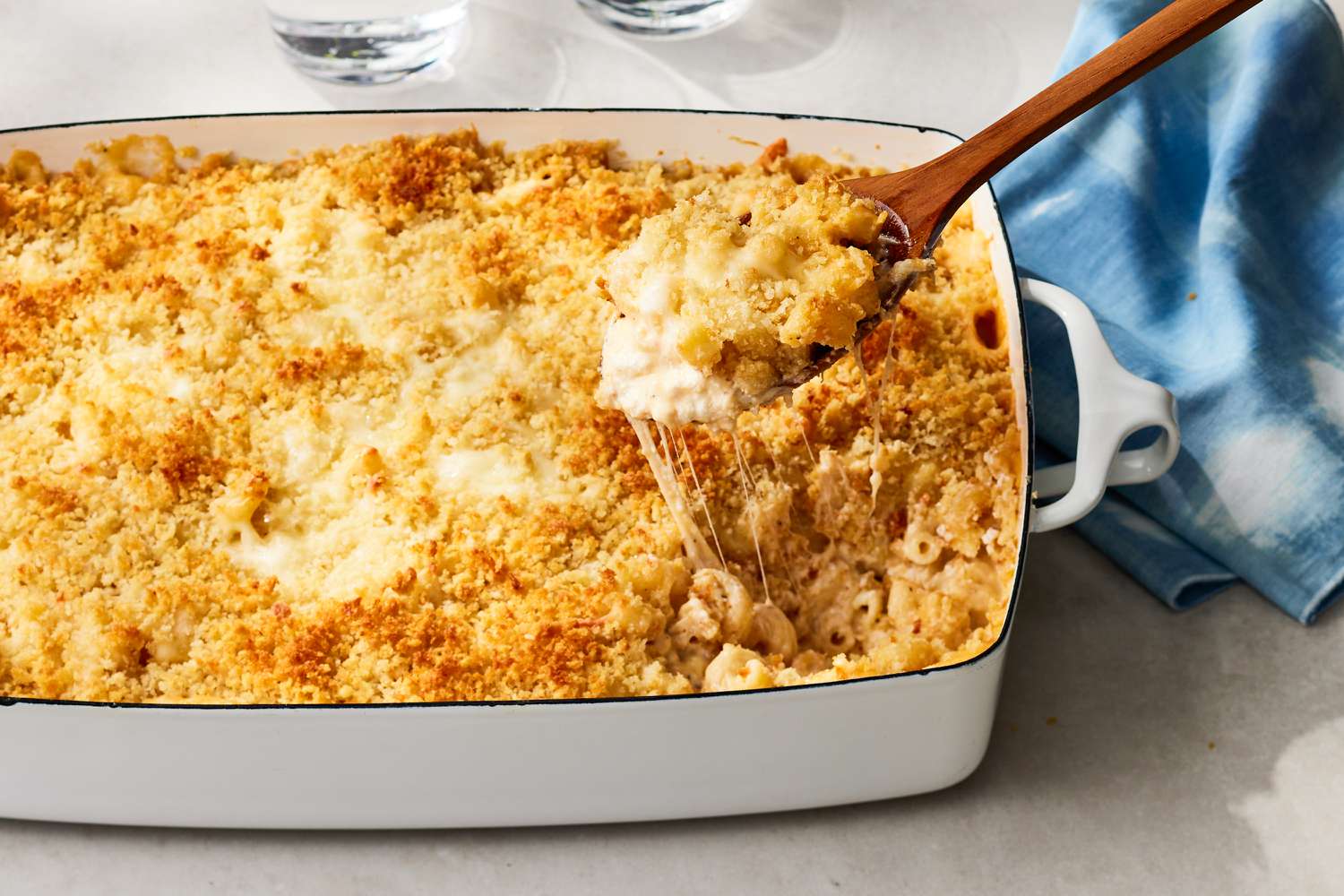 White baking dish of million dollar macaroni and cheese topped with panko with a wooden spoon removing a serving and a blue cloth napkin in the background