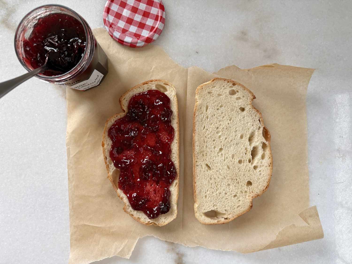 Two slices of bread on parchment paper one spread with jam next to an opened jar of jam with a spoon
