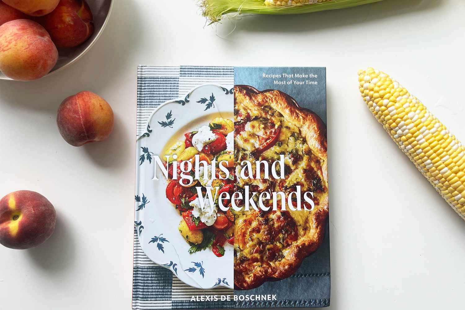 A cookbook titled Nights and Weekends by Alexis de Boschnek surrounded by peaches and corn