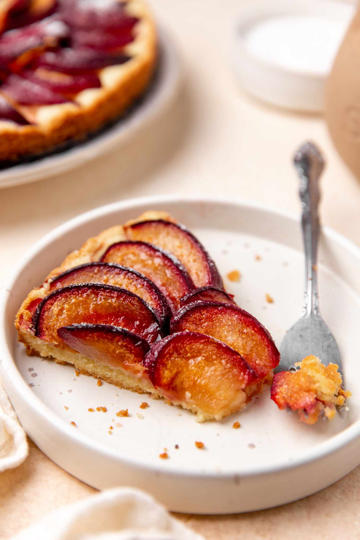 slice of german plum tart on a plate with a fork at a table setting with more tart in the background