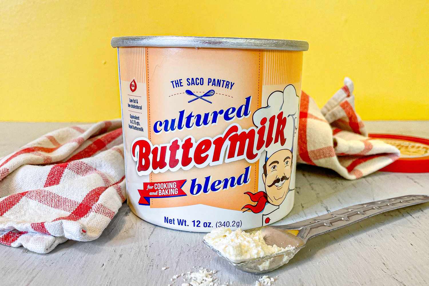 Opened Canister of The Saco Pantry: Cultured Buttermilk Blend on a Table With a Spoonful and a Red and White Checkered Kitchen Towel Next to It