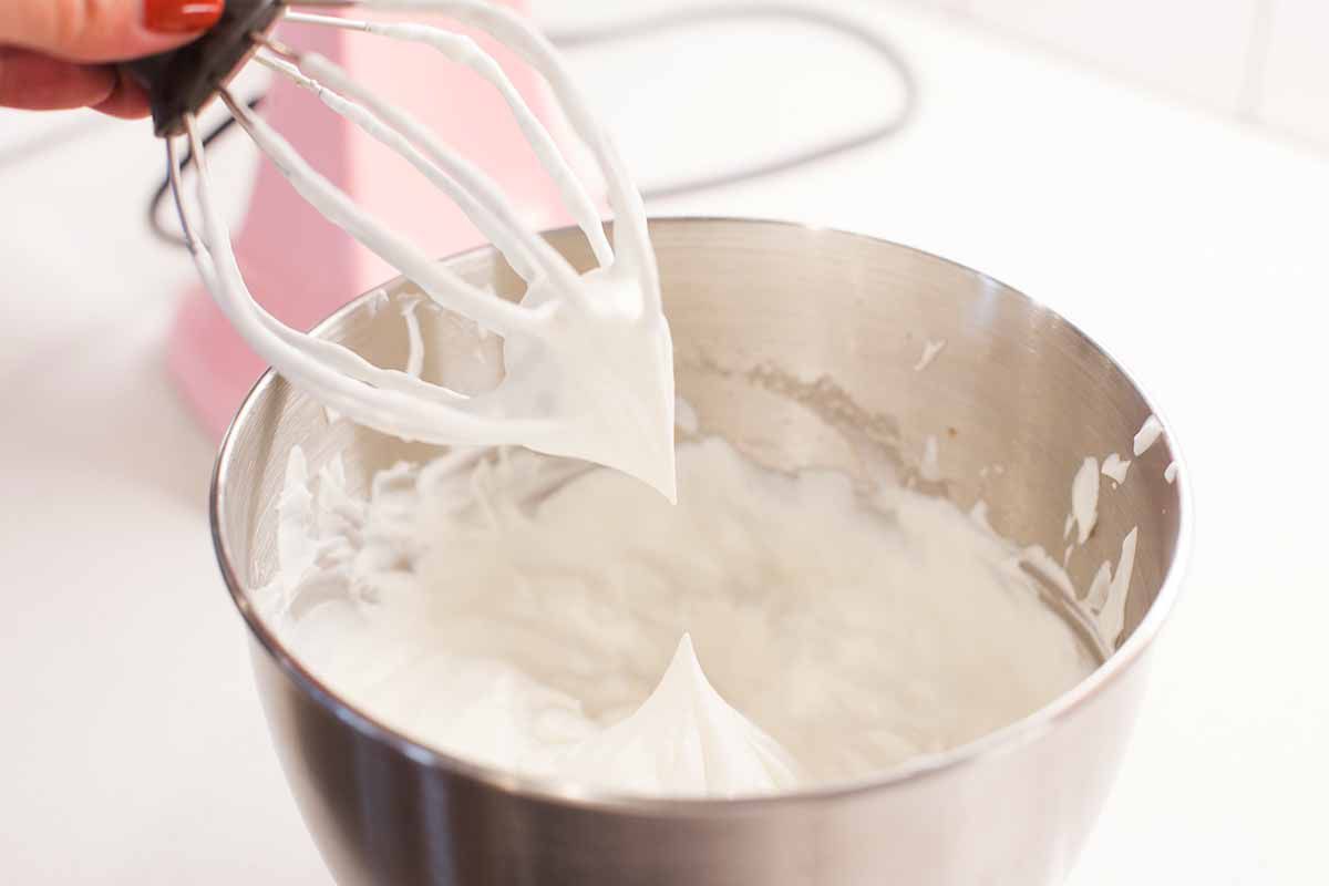 Pink stand mixer with a whisk attachment lifted out of the mixing bowl filled wtih whipped cream.
