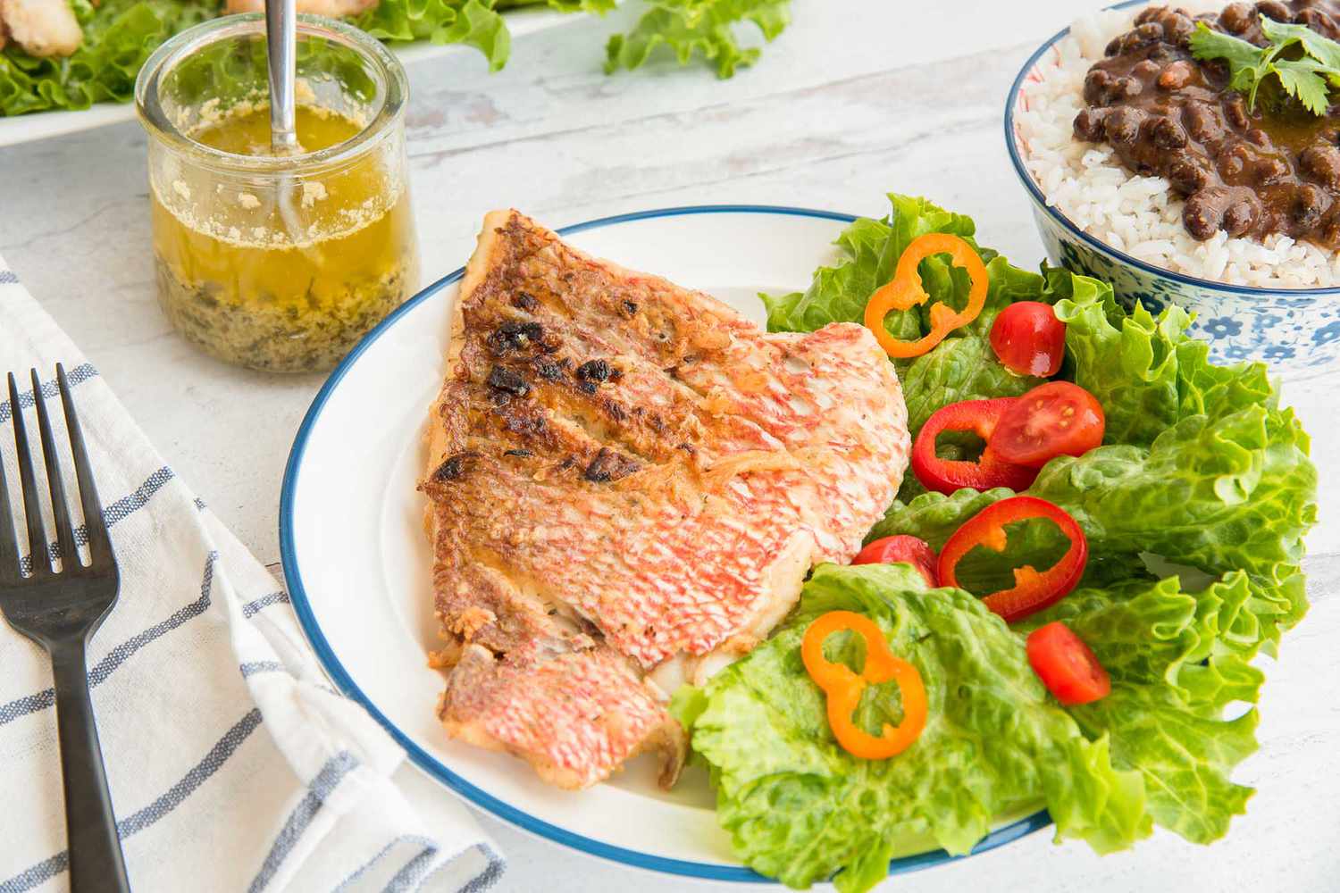 Side view of lenten fish with garlic sauce on a plate with a blue edge. A blue striped napkin and a black fork are to the left of the plate. The large fillet is crispy and pink on the outside. Lettuce and cut red and orange peppers are to the right of the snapper. A bowl of rice and black beans is to the upper right of the image. A jar with a spoon inside has chillo al Ajuillo inside. Behind is a platte with more lettuce and snapper.