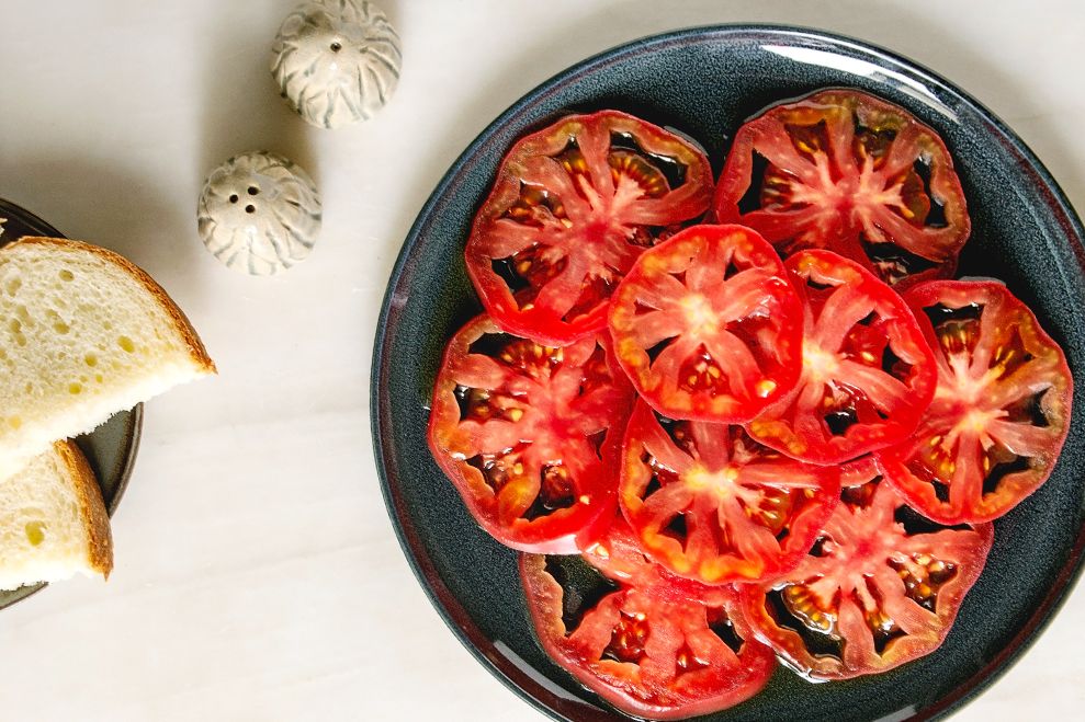 Sliced tomatoes neatly arranged on a dark plate, bread and saltshakers nearby