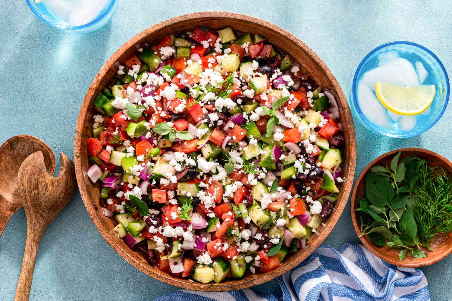 Easy Greek Salad in a Wooden Bowl, and in the Surroundings, Two Glasses of Iced Lemon Water, a Salad Serving Utensils, a Bowl of Herbs, and a White and Blue Striped Kitchen Towel