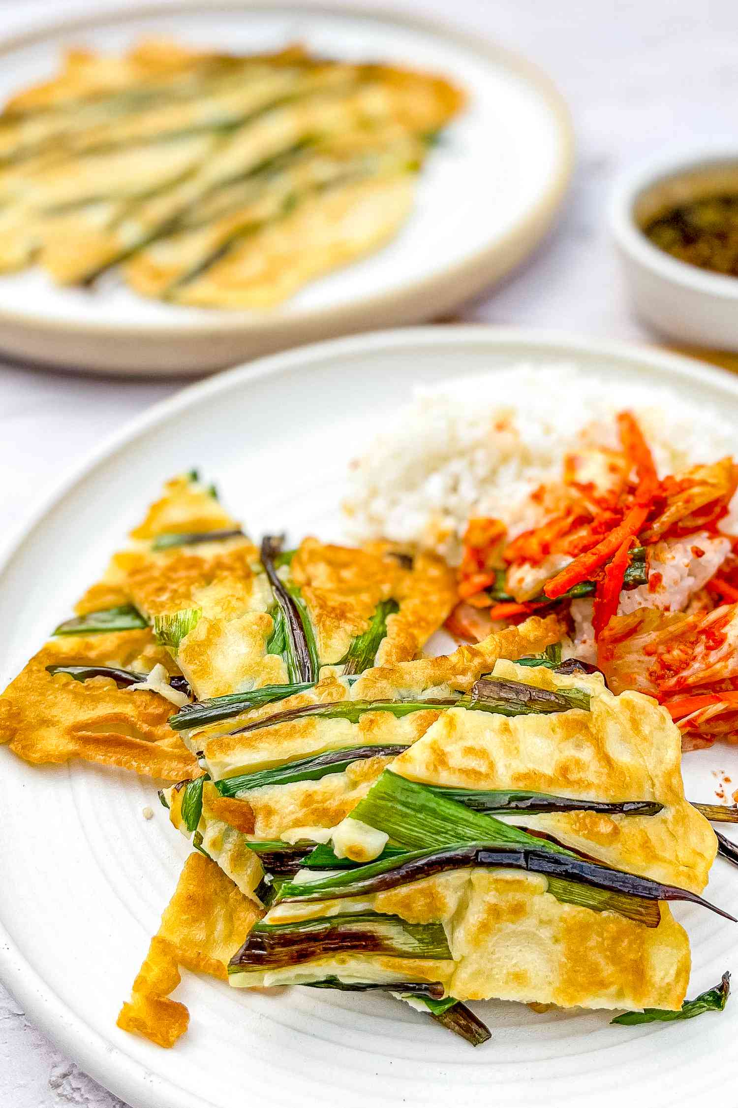 Pajeon (Korean Scallion Pancake), Kimchi, and Rice on a Plate With Another Plate of Pajeon and a Small Bowl of Dipping Sauce in the Background