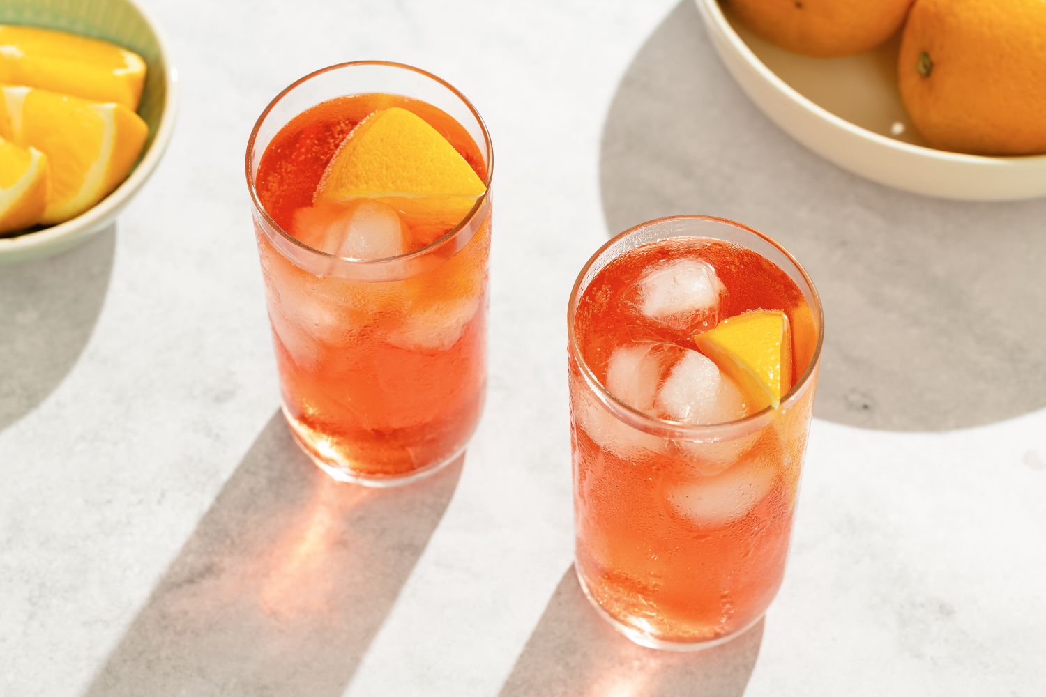 Two glasses of a cocktail with ice and orange slices surrounded by bowls of orange slices and whole oranges on a light surface