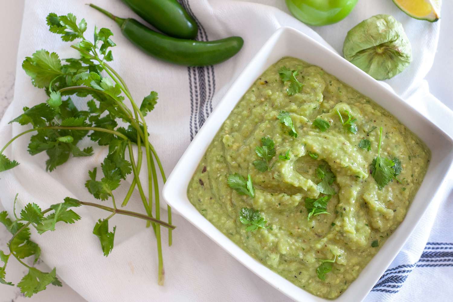 Bowl of Tomatillo Avocado Salsa Surrounded by Tomatillos, Lime Wedges, Cilantro, and Jalapeños