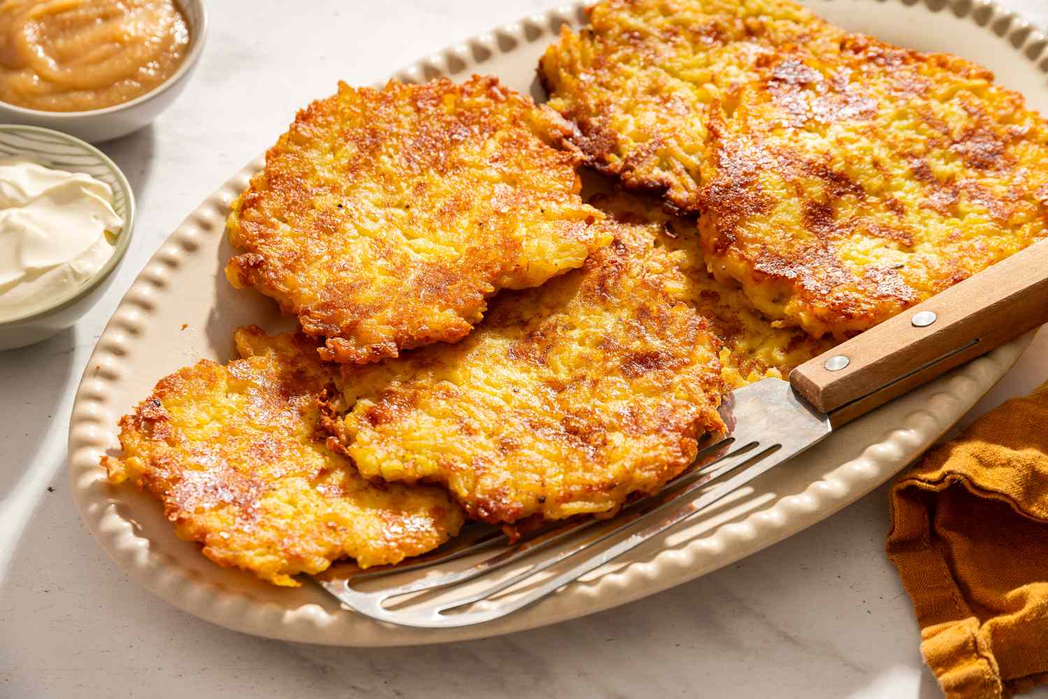 A plate with several crispy latkes a spatula placed beside them bowls of applesauce and sour cream nearby