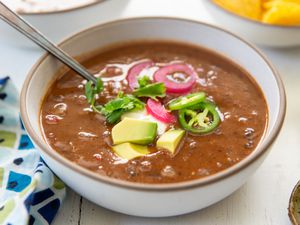 A bowl of black bean soup, topped with avocado cubes, jalapeño slices, pickled onion and and cilantro