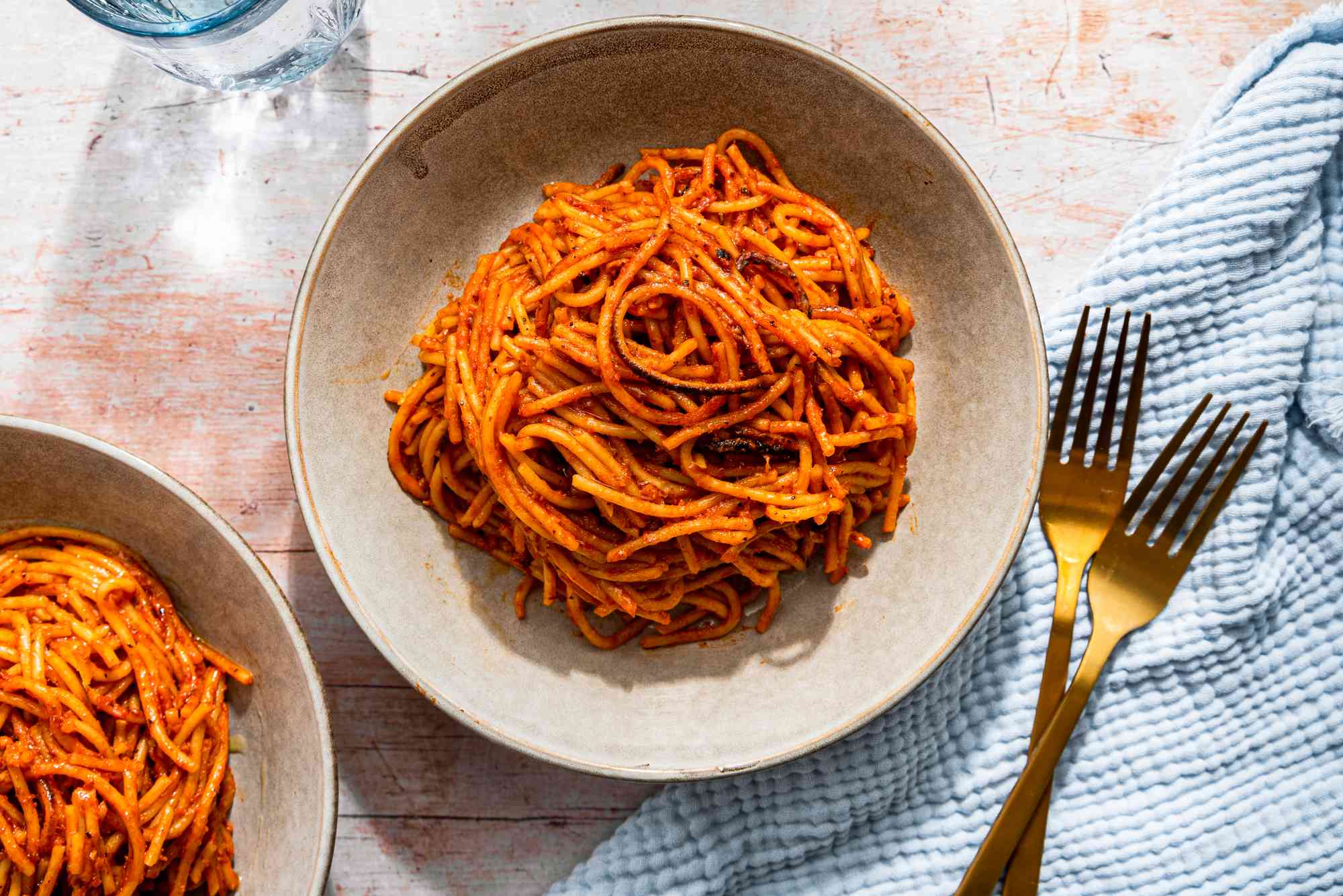 Bowlful of assassin's spaghetti at a table setting with more in a bowl, a glass of water, utensils, and light blue kitchen towel