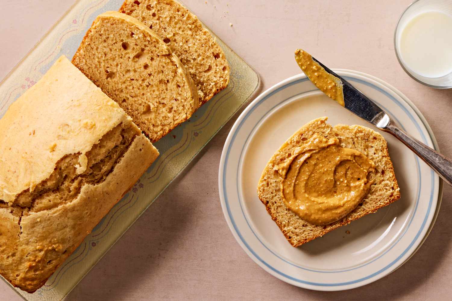 Overhead view of a loaf of retro peanut butter bread next to a plate with a knife and a slice topped with peanut butter next to a glass of milk