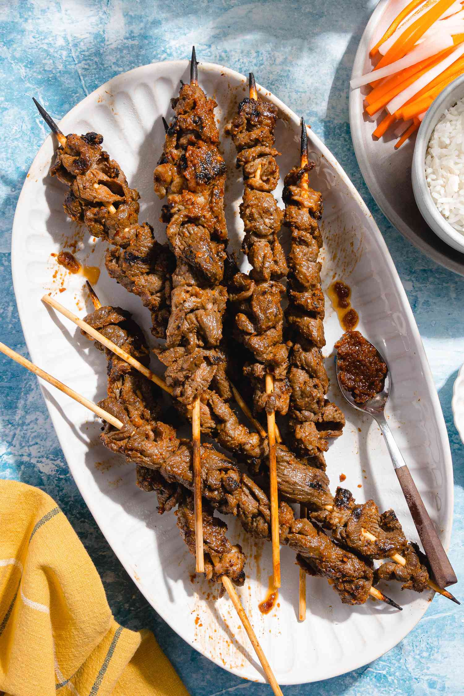 Sach Ko Jakak (Cambodian Grilled Lemongrass Beef Skewers) in a Platter Surrounded by a Plate With Sliced Carrot and Daikon Radish Pickle and a Bowl of Rice and a Yellow Kitchen Towel Next to It