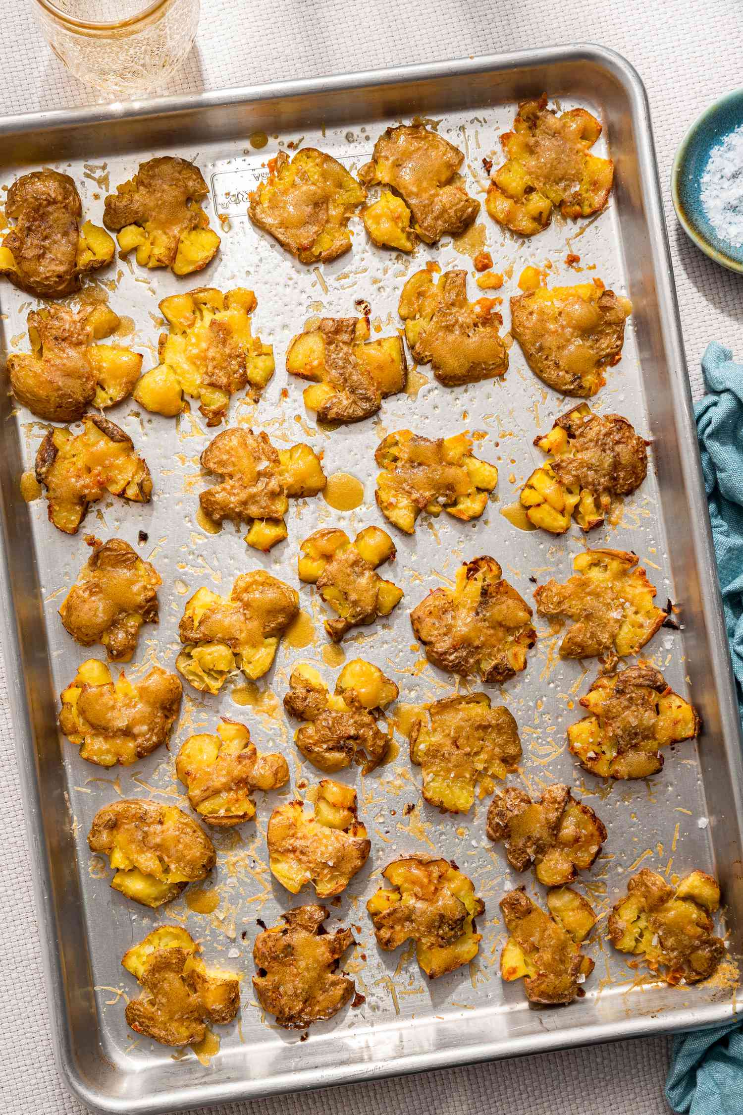 Crash hot potatoes on a baking sheet next to a small saucer with flaky salt, a kitchen towel, and a glass of water