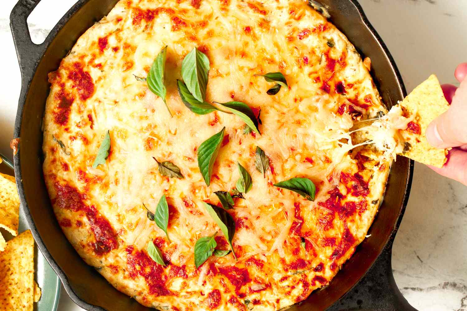 Cast iron skillet with a baked cheese dip garnished with basil and a hand dipping a tortilla chip