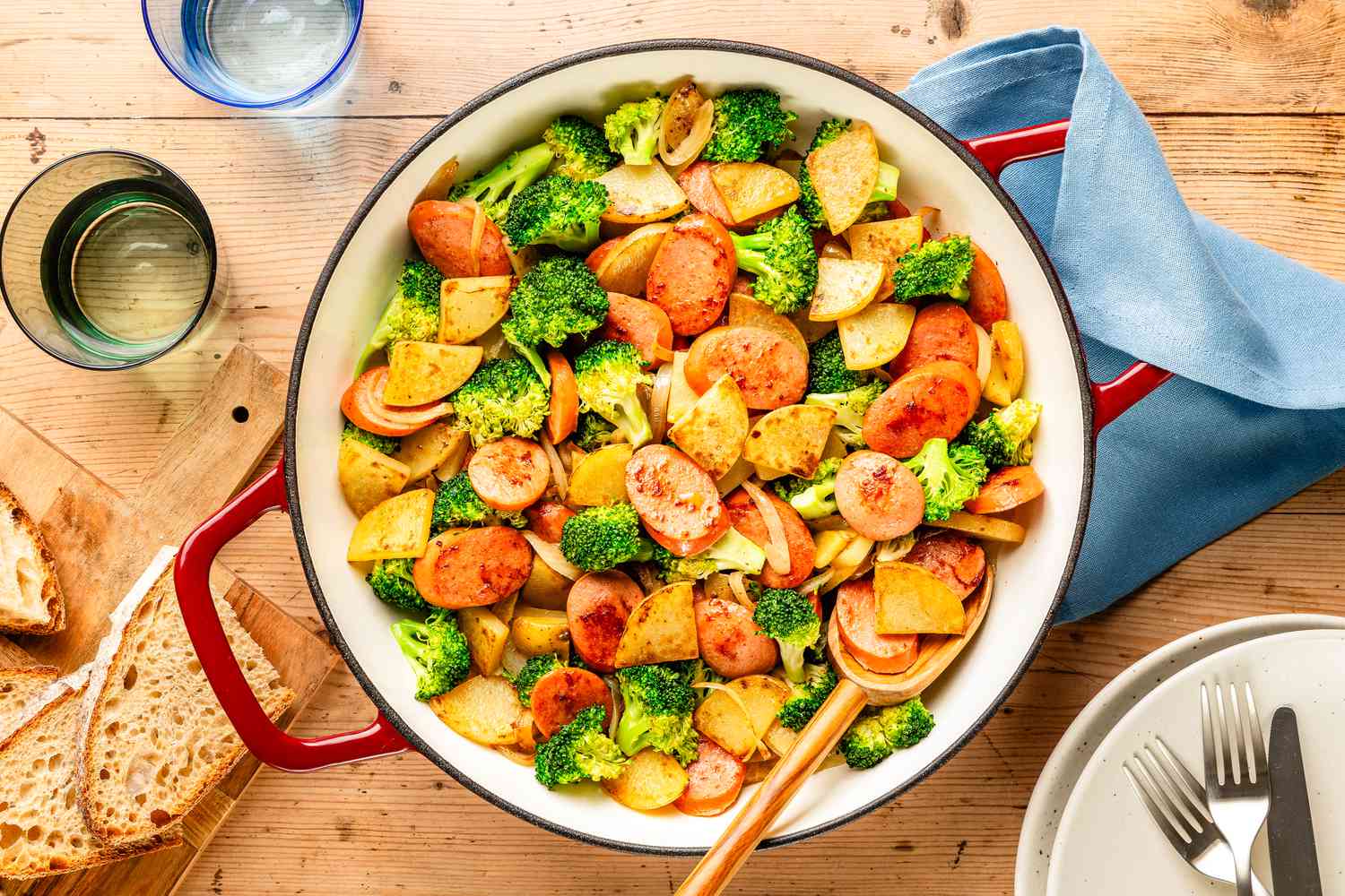 Overhead view of a red and white cast iron skillet of Kielbasa, Potato, and Broccoli Skillet with a serving spoon next to slices of bread on a cutting board, two drinking glasses and a blue fabric napkin