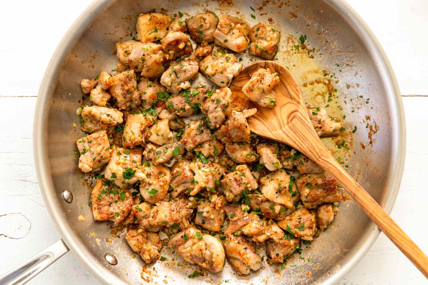 Chunks of cooked chicken in a skillet with herbs and spices, accompanied by a wooden spoon
