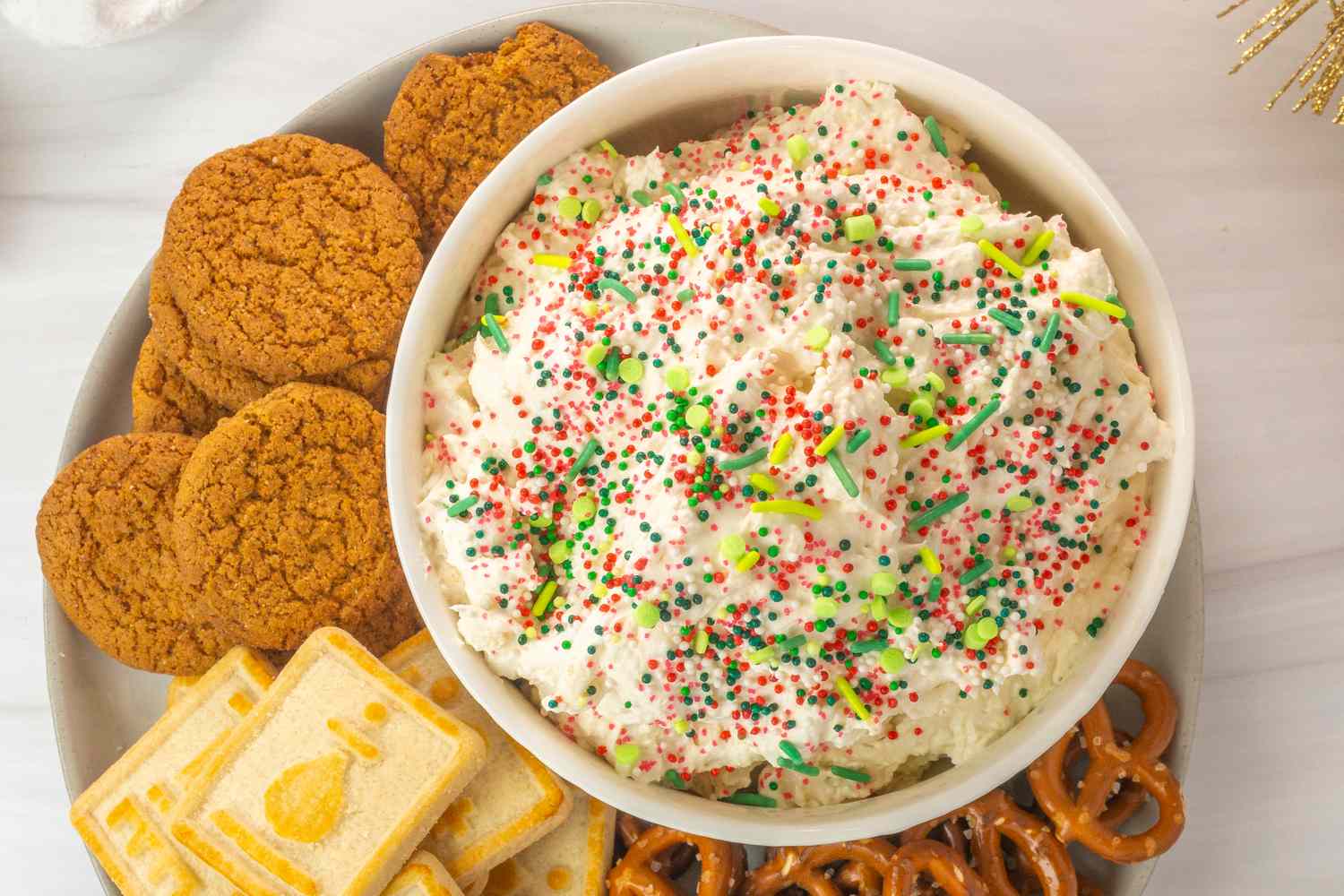 A bowl of dessert dip topped with sprinkles surrounded by cookies and pretzels on a plate