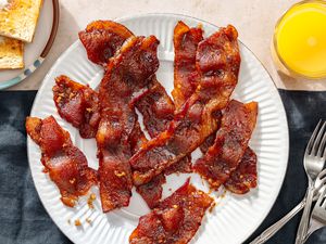 Overhead view of millionaire's bacon on a white plate next to a glass of orange juice, a plate of toast and three forks
