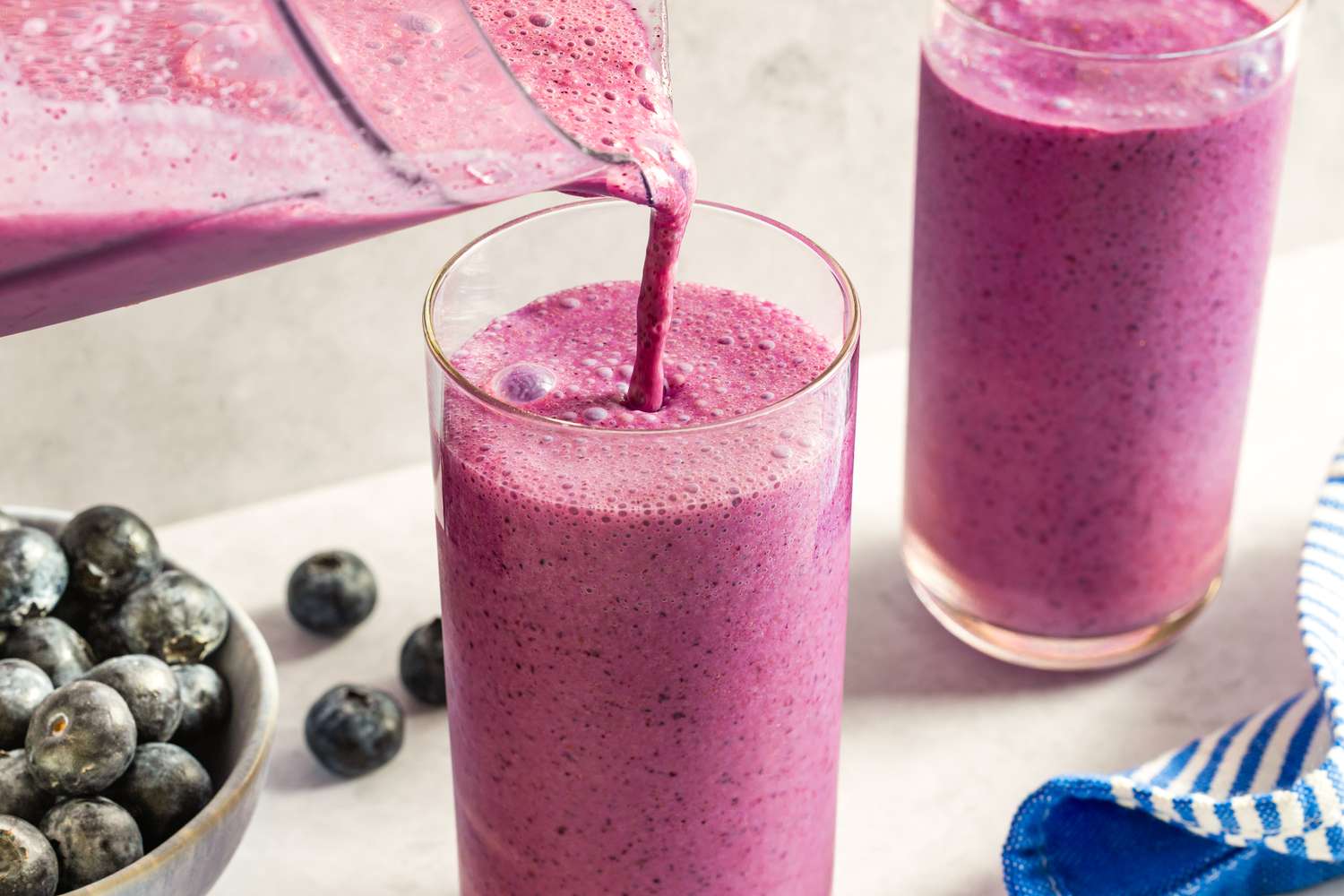 Smoothie being poured into a glass accompanied by a bowl of blueberries on the side