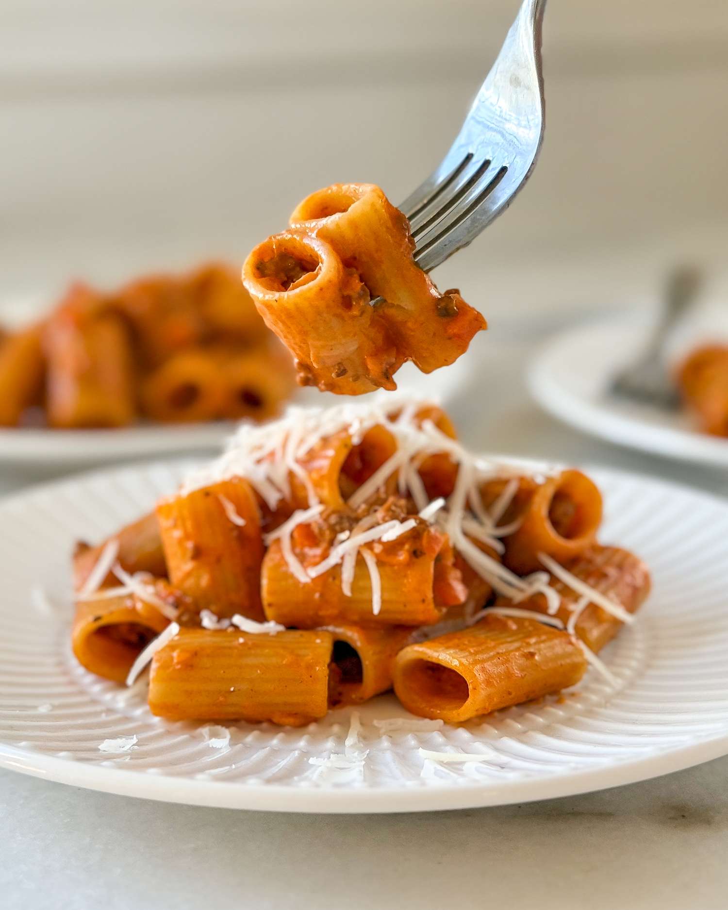 Pasta with a red sauce topped with grated cheese served on a plate with a fork holding up a piece of pasta in focus
