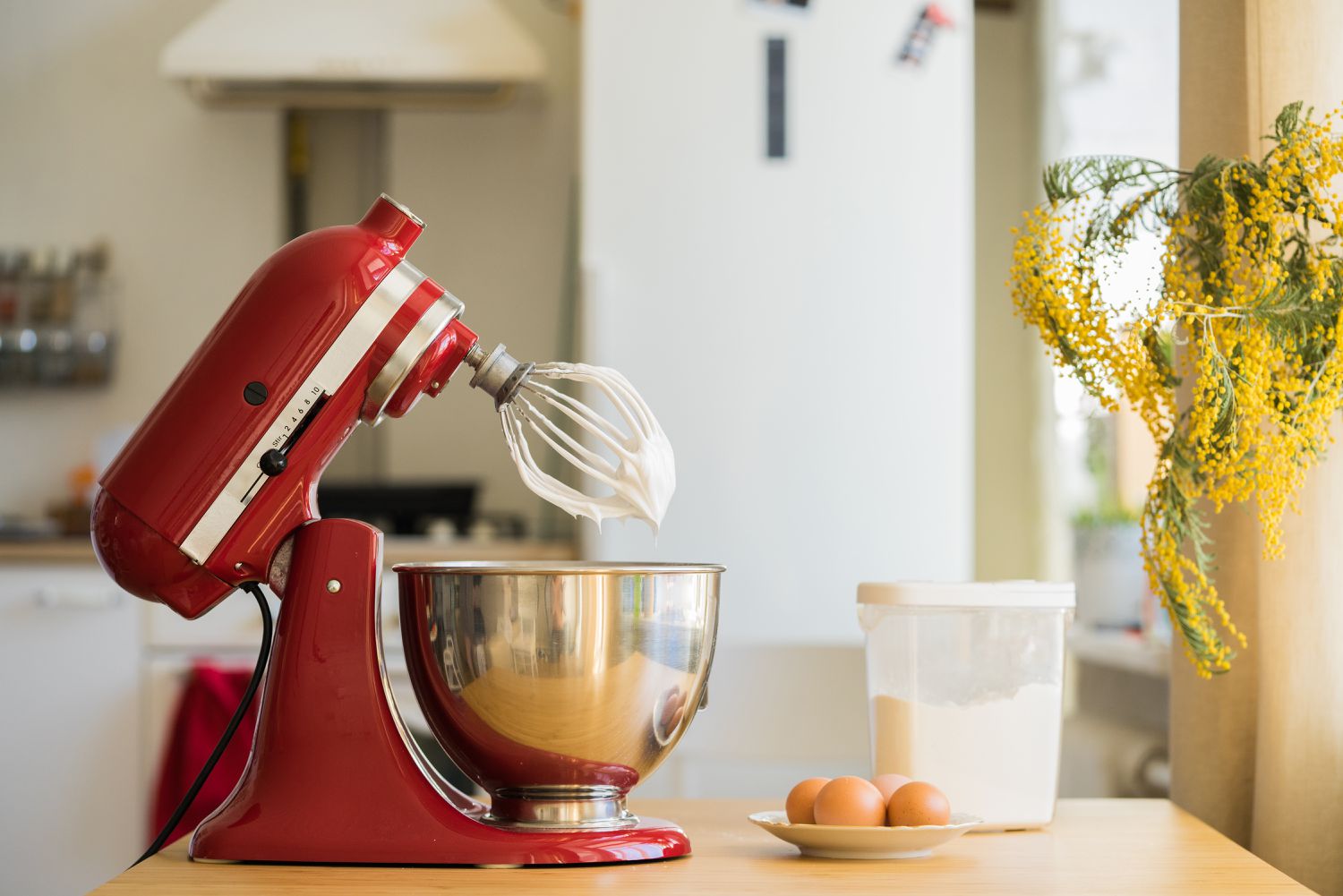 Side view of a red stand mixer and metal bowl with the top section tilted back showing the whisk attachment. The mixer is on a kitchen counter next to a plate of eggs and container of flour